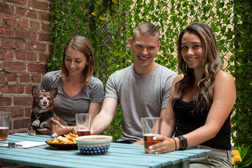 three people with a dog on a picnic table with drinks