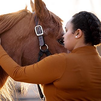 woman with braided hair and an orange-brown shirt in front of a brown horses head